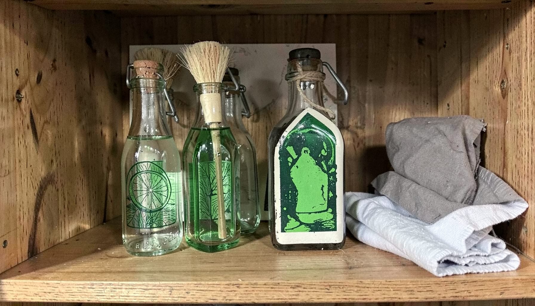 Flacons en verre de vinaigre blanc et savon noir, brosses en fibres naturelles et chiffons en coton, sur une étagère en bois
