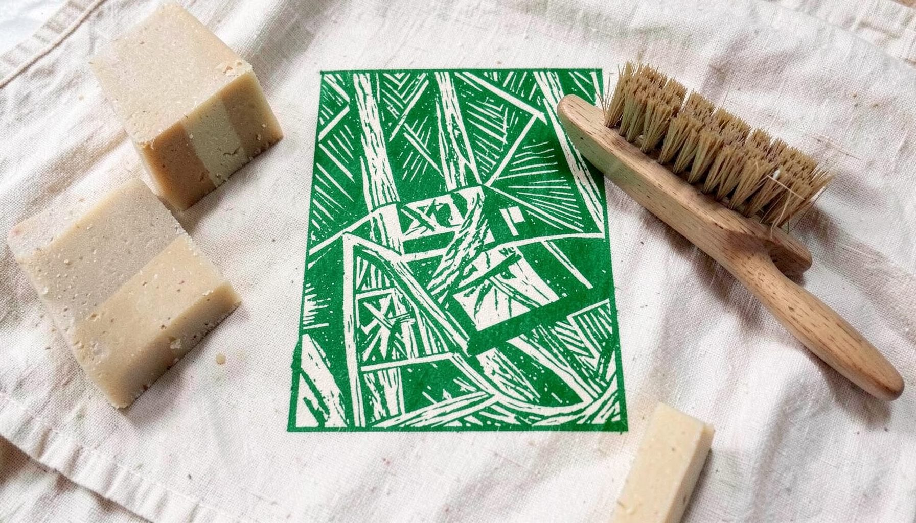 Savons solides, shampoings en barre et brosses en bois posés sur un linge en coton écru, tons beige, vert sauge et blanc